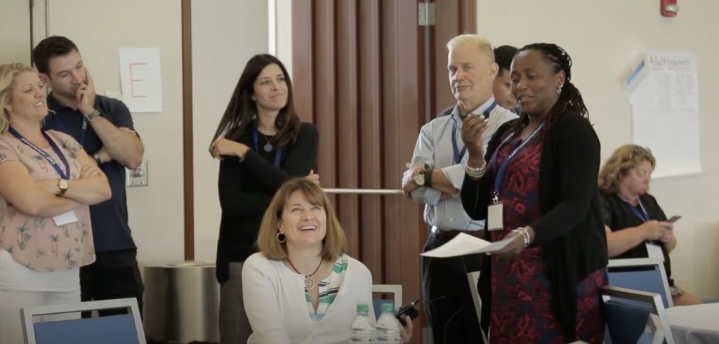 A snapshot of a diverse group of six attendees interacting during a networking session at a previous annual CAST UDL symposium. The group comprises three White women, a Black woman holding a worksheet, and two white men. Source: YouTube/CAST 