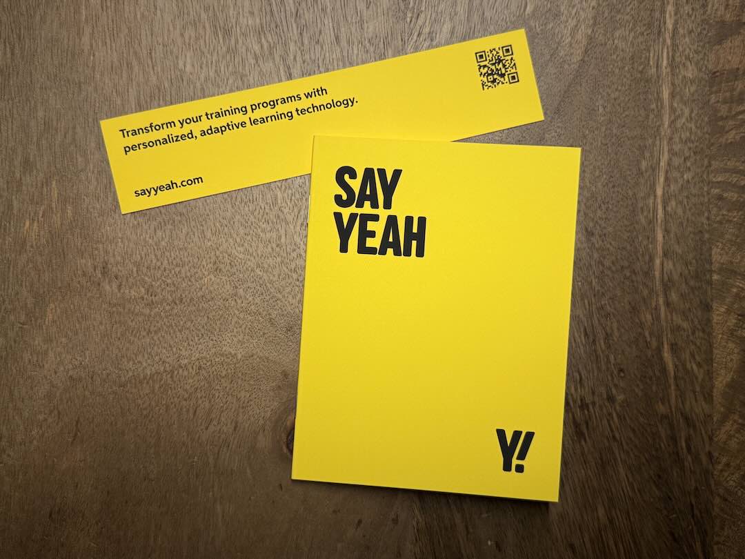 Say Yeah's sleek crisp yellow pocket notebook with a bold "Say Yeah" and accompanying "Y!" logo sits alongside a bookmark which reads "Transform your training programs with personalized, adaptive learning technology." with a QR code for sayyeah.com.